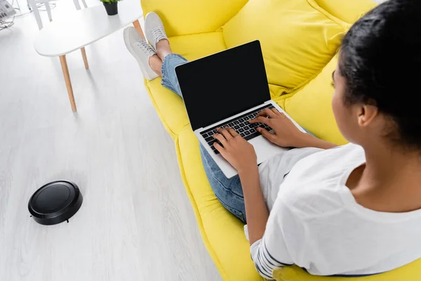 Unscharfe Afroamerikanerin benutzt Laptop in der Nähe von Staubsaugerroboter — Stockfoto