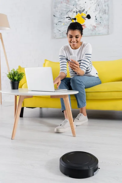 Robotic vacuum cleaner near blurred african american freelancer with smartphone and laptop — Stock Photo
