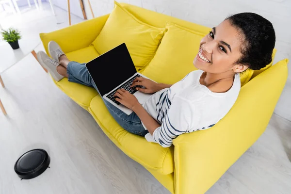 Blick aus der Vogelperspektive auf afrikanische Freiberufler mit Laptop auf Couch in der Nähe von Staubsaugerrobotern — Stockfoto