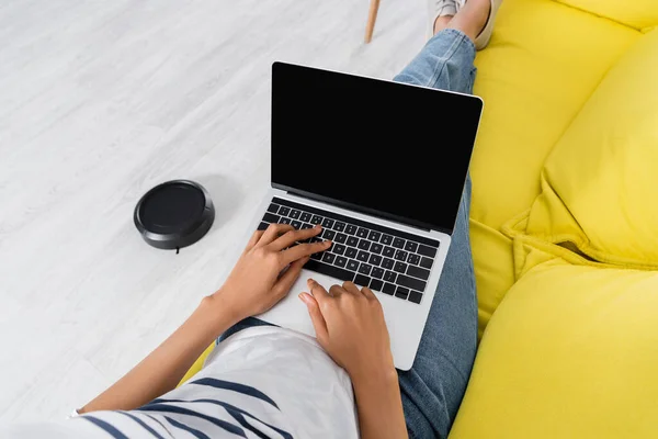 Ausgeschnittene Ansicht einer afrikanisch-amerikanischen Frau mit Laptop mit leerem Bildschirm in der Nähe von Staubsaugerrobotern — Stockfoto