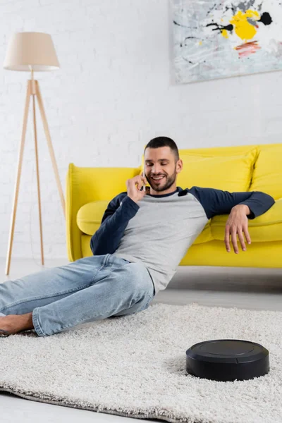Роботизированный пылесос на ковре рядом с улыбающимся человеком разговаривающим по смартфону на размытом фоне — стоковое фото