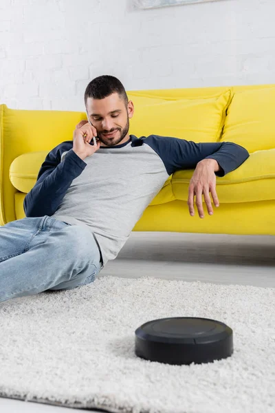 Hombre joven hablando en el teléfono inteligente cerca borrosa aspiradora robótica - foto de stock