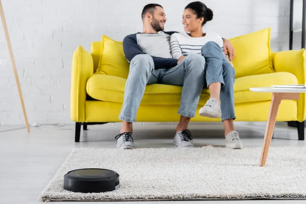 Robotic vacuum cleaner near blurred interracial couple on couch — Stock Photo