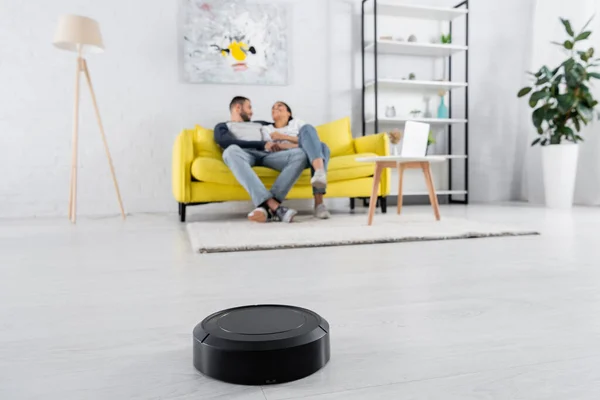 Robotic vacuum cleaner on floor near blurred couple in living room — Stock Photo