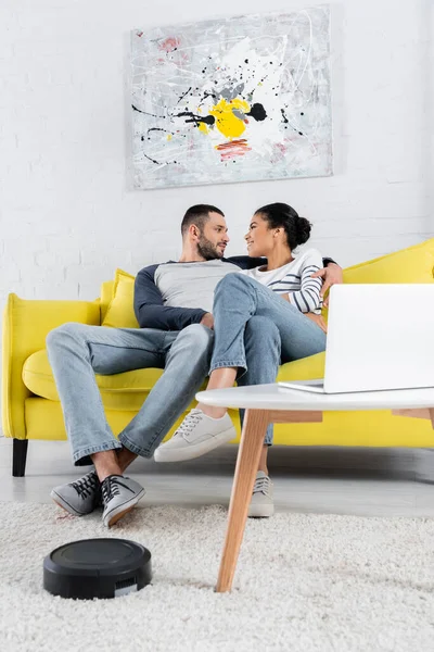 Side view of man hugging african american girlfriend near blurred laptop and robotic vacuum cleaner — Stock Photo