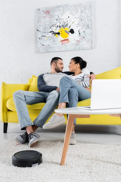 Vista lateral del hombre abrazando a la novia afroamericana cerca de la computadora portátil borrosa y la aspiradora robótica - foto de stock