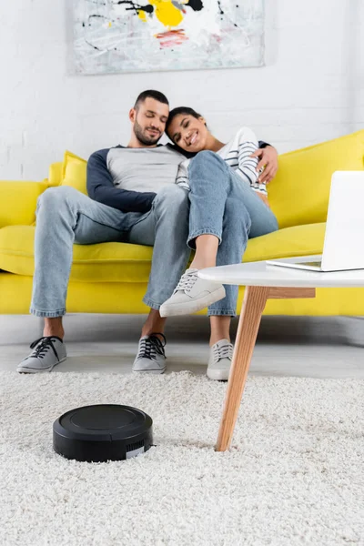 Aspirador robótico perto de laptop e casal multiétnico sorridente no sofá — Fotografia de Stock