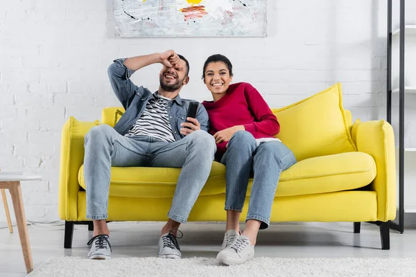 Positive multiethnic couple with remote controller sitting on couch — Stock Photo