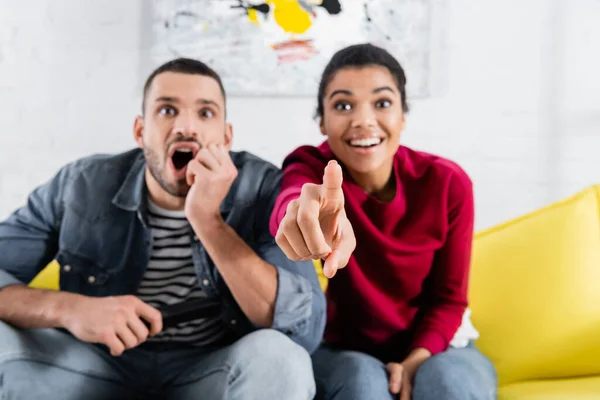 Mujer afroamericana borrosa apuntando con el dedo cerca de novio sorprendido con controlador remoto - foto de stock