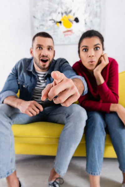 Hombre borroso señalando con el dedo a la cámara cerca de la novia afroamericana en casa - foto de stock