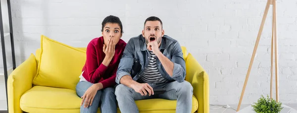 Pareja asombrada con mando a distancia sentado en el sofá, pancarta — Stock Photo