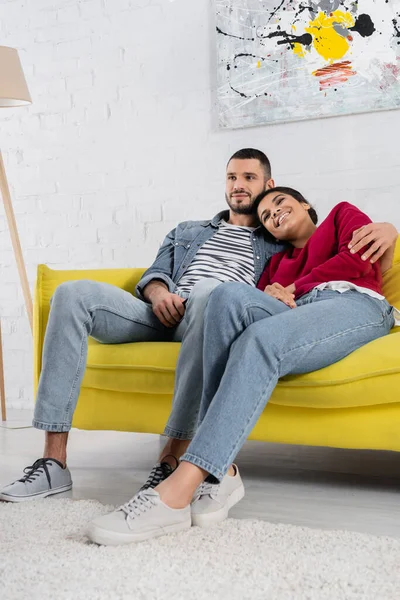 Joven abrazando sonriente afro-americana novia en sofá - foto de stock