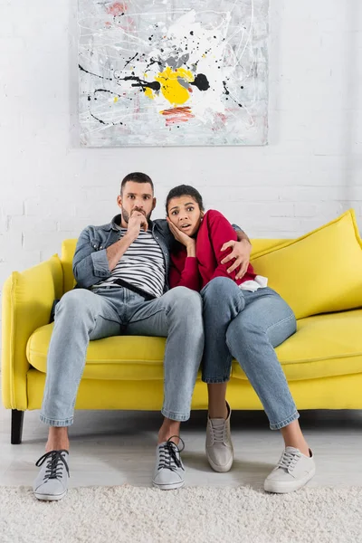 Scared interracial couple looking at camera near remote controller on couch — Stock Photo