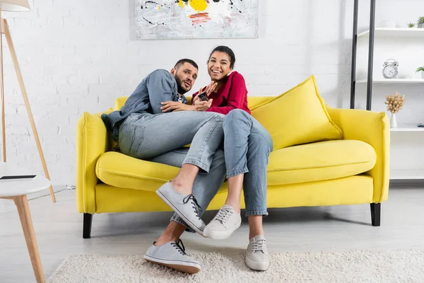 Scared man sitting near cheerful african american girlfriend with remote controller — Stock Photo