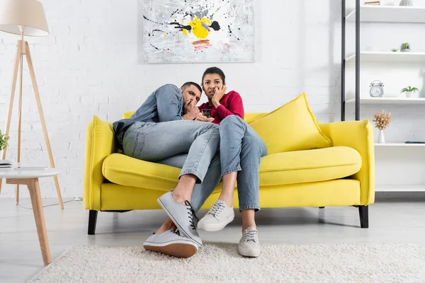 Scared african american woman holding remote controller near boyfriend in living room — Stock Photo