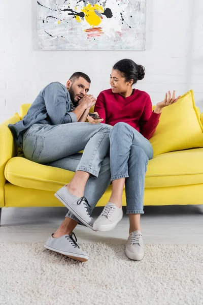 Femme afro-américaine souriante avec télécommande regardant petit ami effrayé dans le salon — Photo de stock