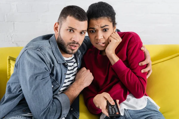 Scared man hugging african american girlfriend with remote controller — Stock Photo