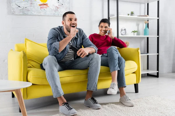 Cheerful man holding remote controller near smiling girlfriend talking on smartphone on couch — Stock Photo