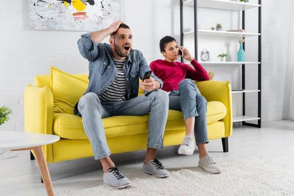 Emocionado hombre con mando a distancia viendo la televisión cerca escéptica novia afroamericana hablando en el teléfono inteligente - foto de stock