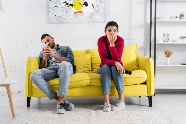 Verängstigte Afroamerikanerin hält Fernbedienung in der Nähe ihres Freundes mit Smartphone auf Couch — Stockfoto