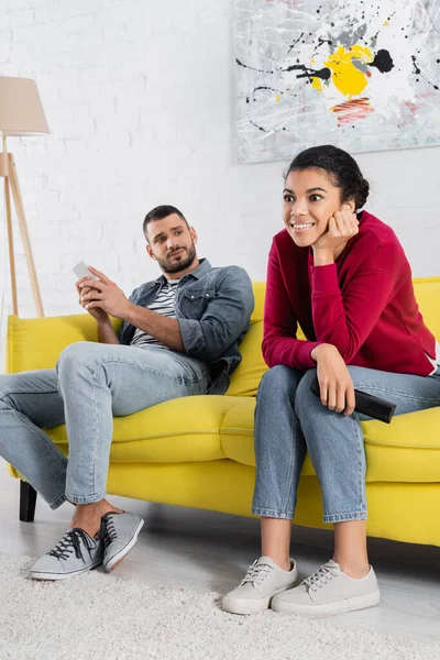 Sonriente mujer afroamericana con controlador remoto sentado cerca de novio con teléfono inteligente en el sofá - foto de stock
