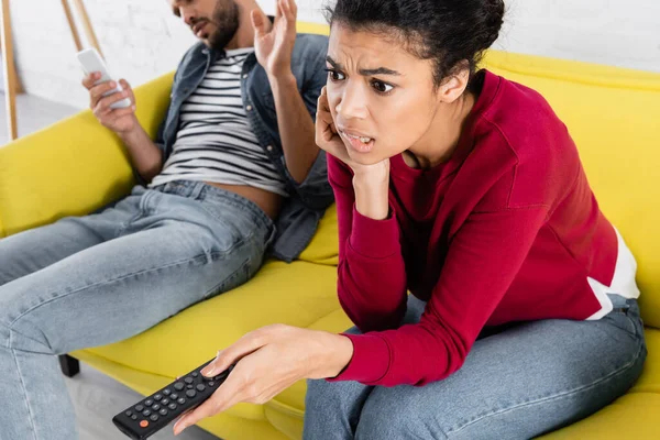 Preoccupata donna afro-americana con telecomando vicino al fidanzato con smartphone a casa — Foto stock
