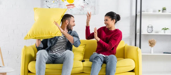 Vista lateral de la pareja interracial positiva almohada luchando en casa, pancarta - foto de stock