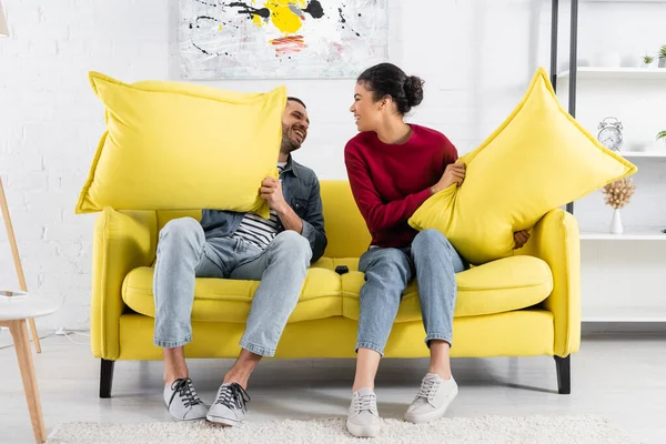 Alegre pareja multiétnica sosteniendo almohadas en el sofá - foto de stock