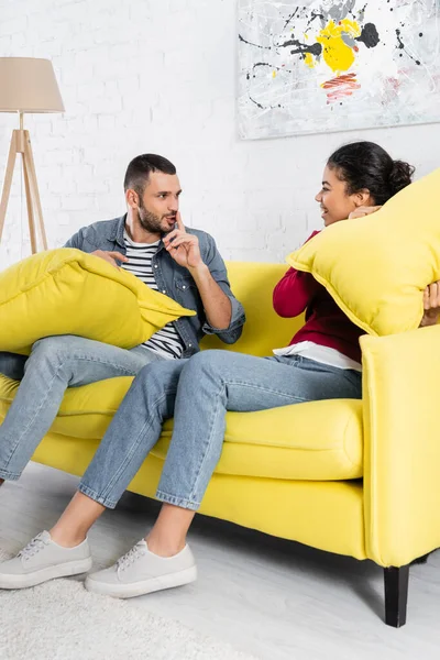 Man showing shh gesture at african american girlfriend with pillow — Stock Photo