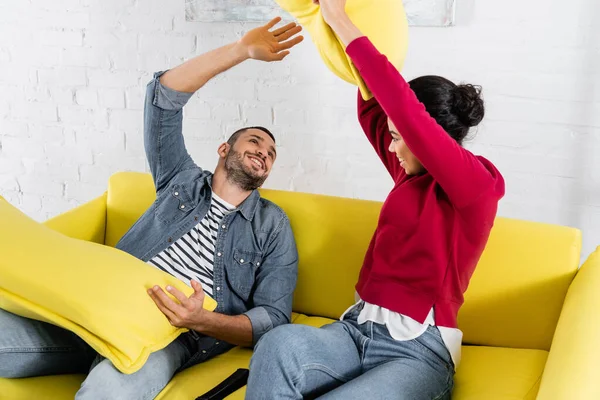 Casal multiétnico alegre travesseiro lutando perto de controlador remoto no sofá — Fotografia de Stock