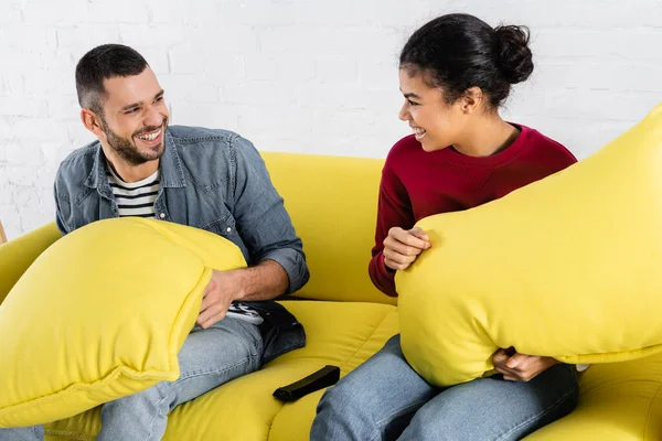 Feliz interracial casal segurando travesseiros perto de controle remoto no sofá — Fotografia de Stock