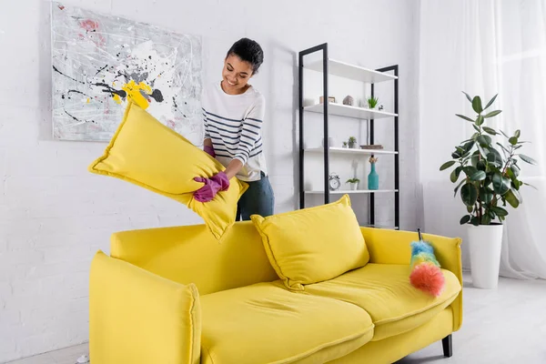 Femme afro-américaine souriante tenant l'oreiller du canapé près de la brosse à poussière à la maison — Photo de stock