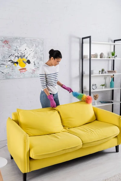Side view of smiling african american woman in rubber gloves holding dust brush while cleaning couch — Stock Photo