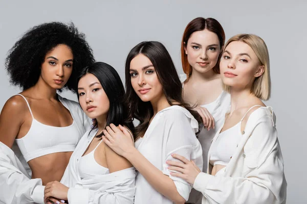 Modelos americanos asiáticos y africanos en camisas blancas posando con mujeres jóvenes aisladas en gris - foto de stock