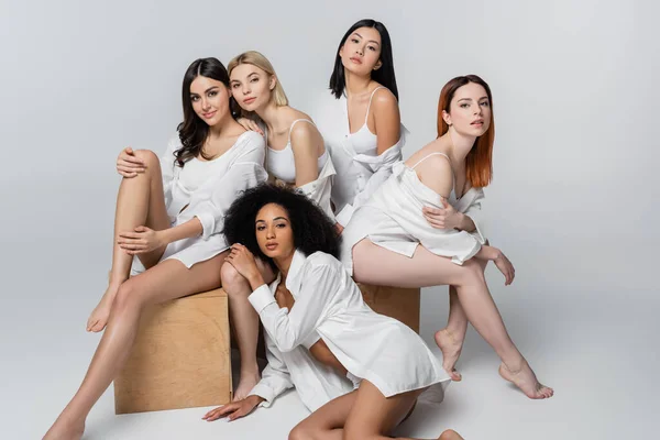 Brunette asian and african american models in white shirts posing near wooden cubes with young women on grey — Stock Photo