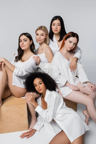 Brunette asian and african american women in white shirts posing near wooden cubes with young models on grey — Stock Photo