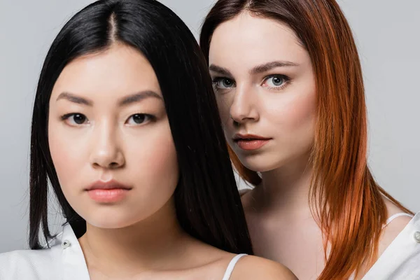 Redhead woman looking at camera near blurred asian model isolated on grey — Stock Photo
