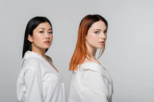 Young and brunette asian woman near redhead model looking at camera isolated on grey — Stock Photo