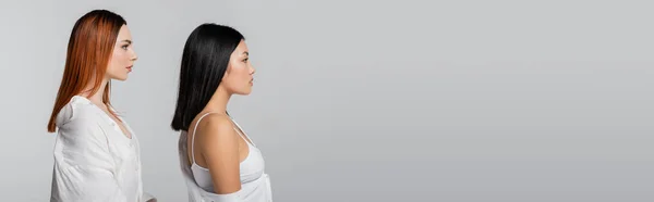 Side view of brunette asian woman and redhead model posing isolated on grey, banner — Stock Photo