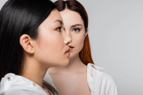 Rousse femme posant près floue et brunette asiatique modèle isolé sur gris — Photo de stock