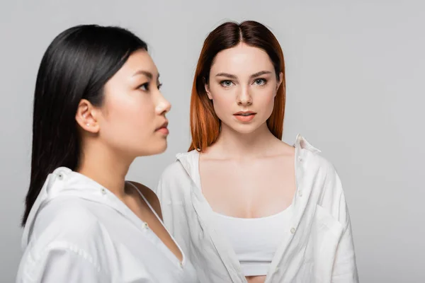 Young redhead woman posing near blurred and brunette asian model isolated on grey — Stock Photo