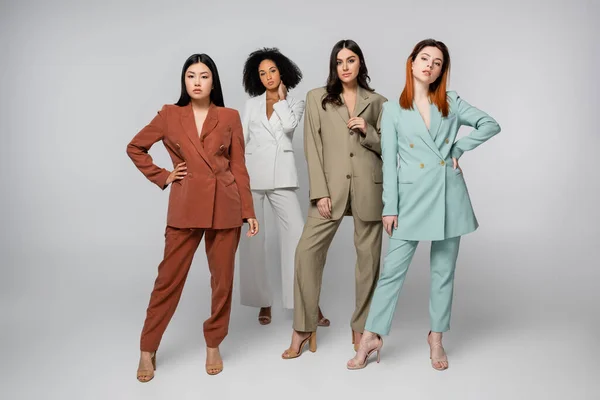 Full length of multiethnic models in pastel color suits posing on grey — Stock Photo