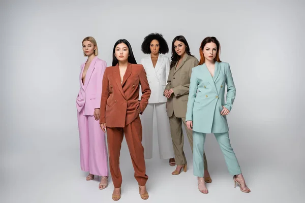 Full length of interracial models in stylish suits posing with hands in pockets on grey — Stock Photo