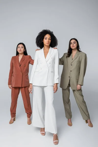 Full length of interracial women in stylish suits standing on grey — Stock Photo