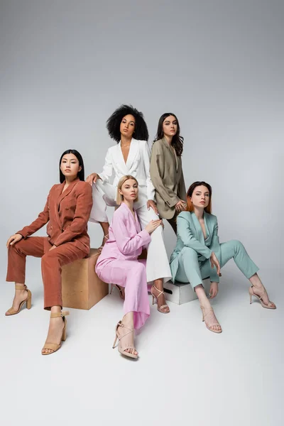 Full length of interracial models in stylish pastel suits sitting while posing on grey — Stock Photo
