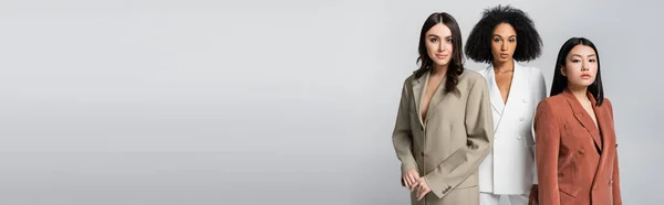 Hübsche interrassische Frauen in trendigen Anzügen posieren isoliert auf grauem Banner — Stockfoto