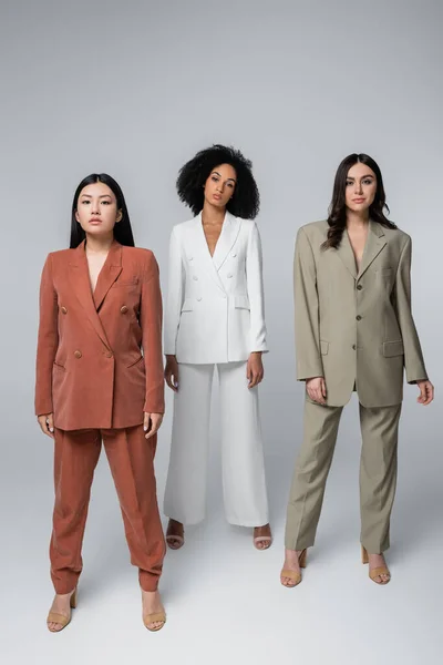 Full length of pretty interracial women in trendy suits posing on grey — Stock Photo