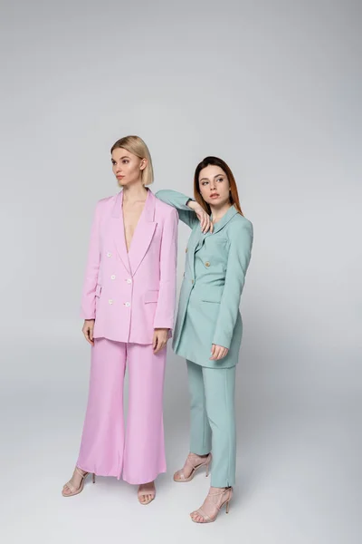 Full length of blonde and redhead women in pink and blue pastel suits posing on grey — Stock Photo