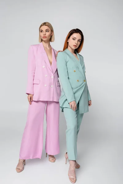 Full length of young blonde and redhead models in pink and blue pastel suits posing on grey — Stock Photo