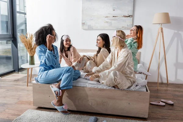 Group of cheerful interracial friends in pajamas sitting on bed during slumber party — Stock Photo